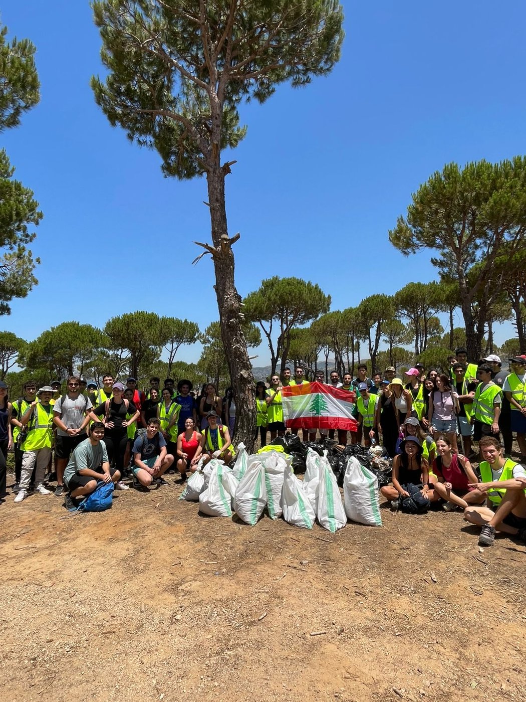 EcoCitizens volunteers in safety vests gathered under pine trees with a Lebanese flag after a cleanup